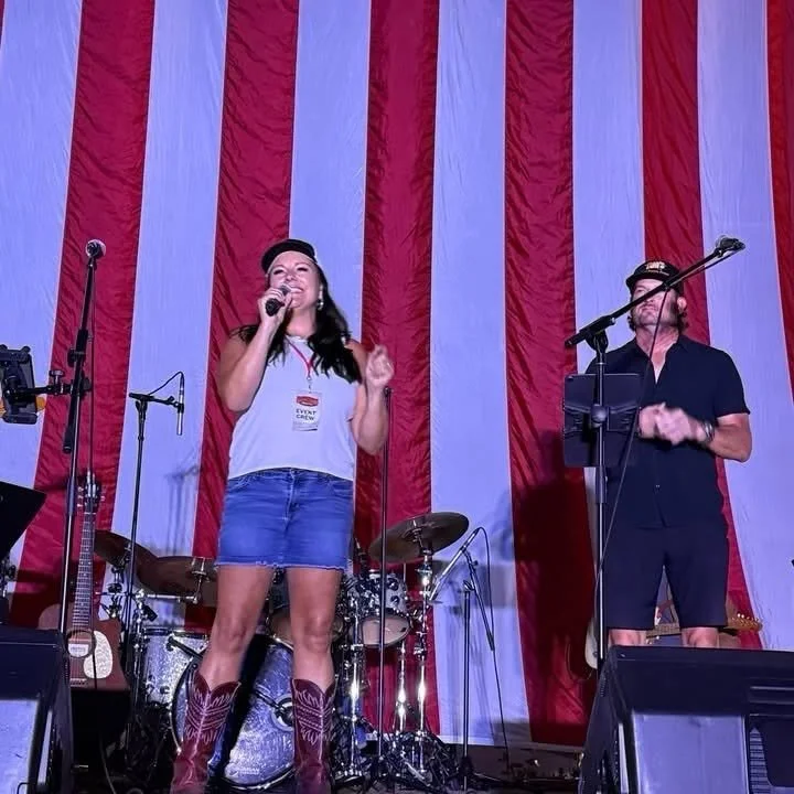 A woman and man on stage with microphones, set against a red and white striped curtain, possibly at a patriotic or country-themed event, with musical instruments behind them.