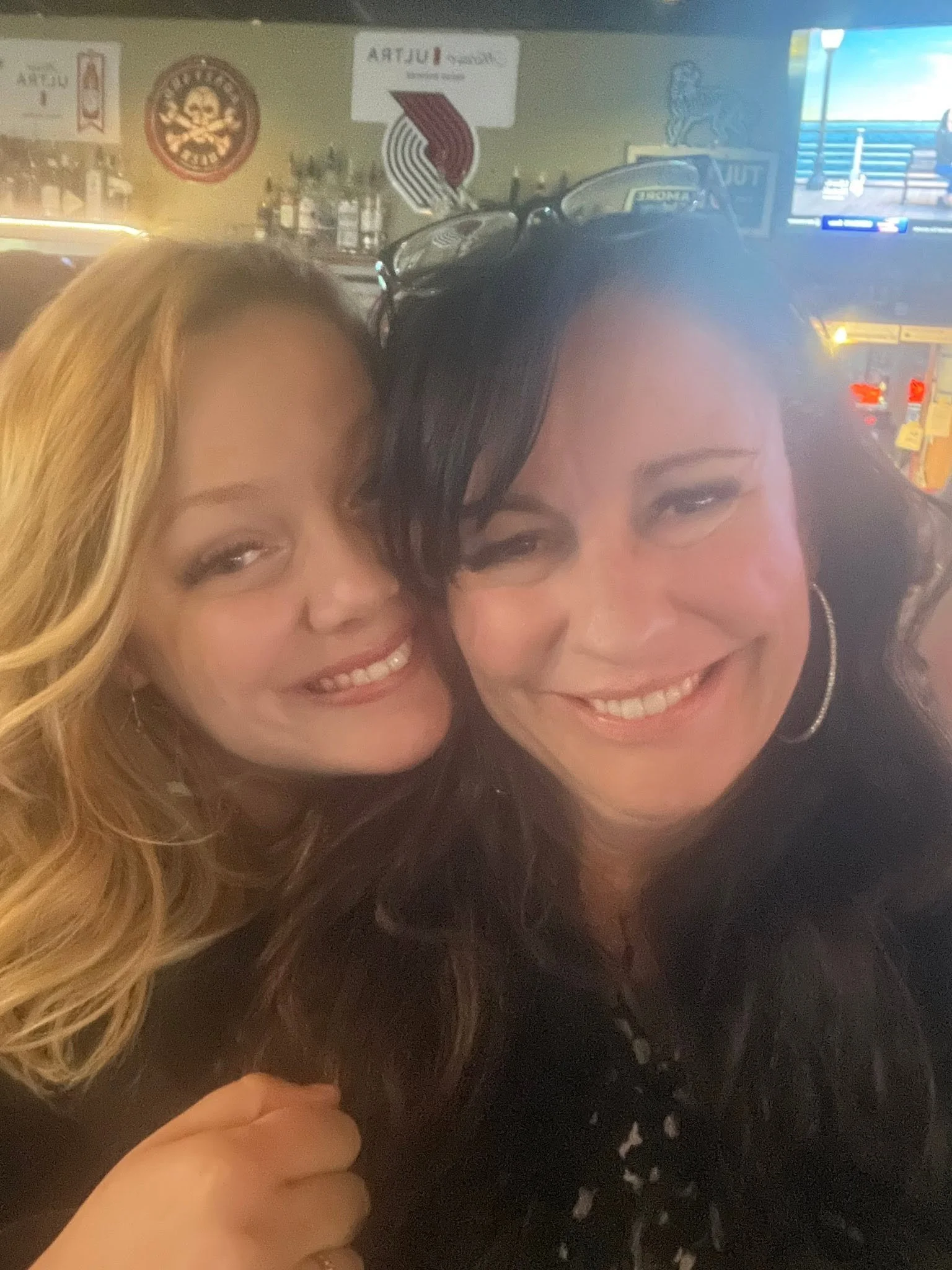 Two women smiling close together in a bar or restaurant, with a bar and TV in the background.