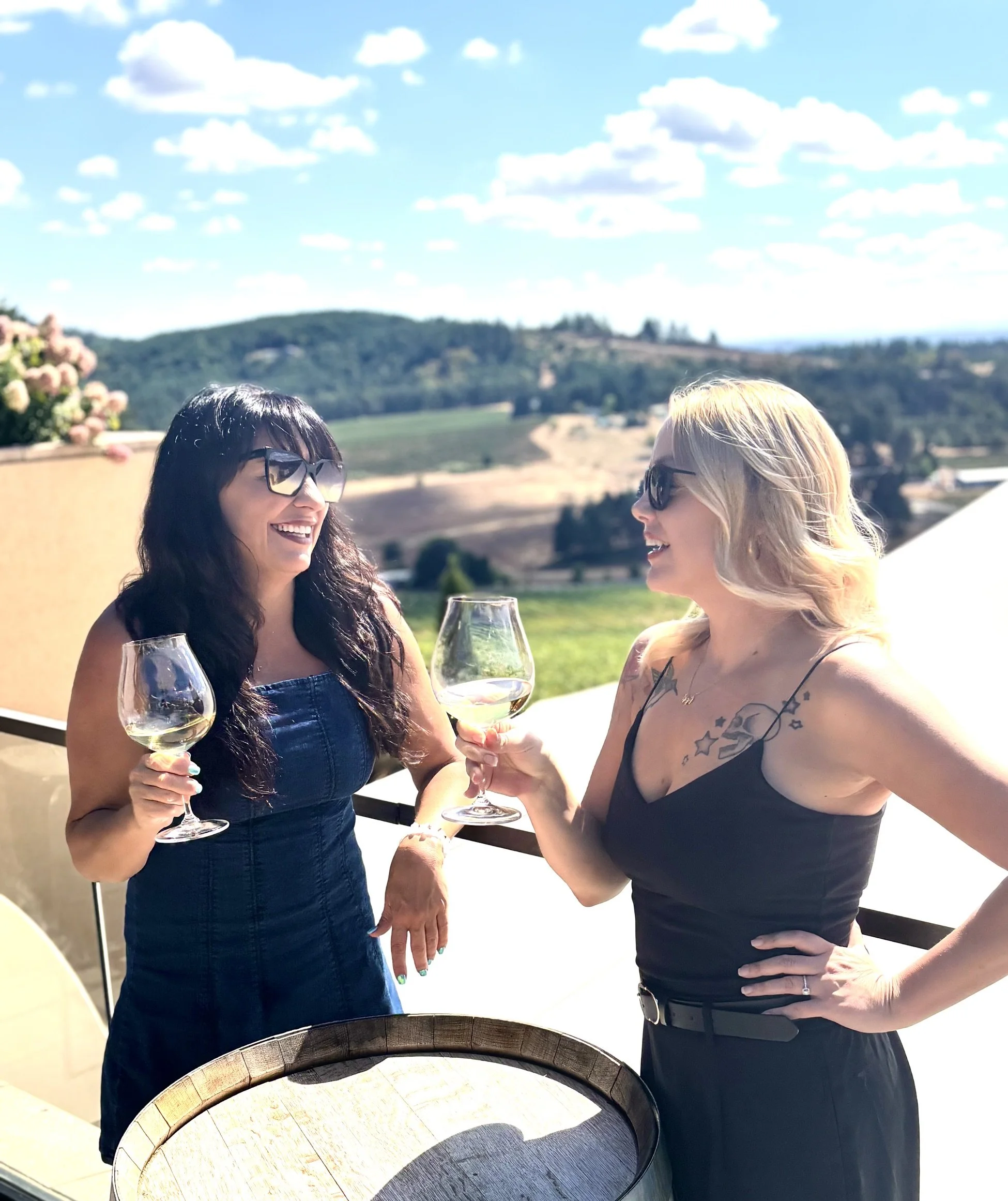 Two women smiling and wearing sunglasses, holding glasses of white wine, enjoying an outdoor setting on a sunny day with a scenic background of rolling hills and blue sky.