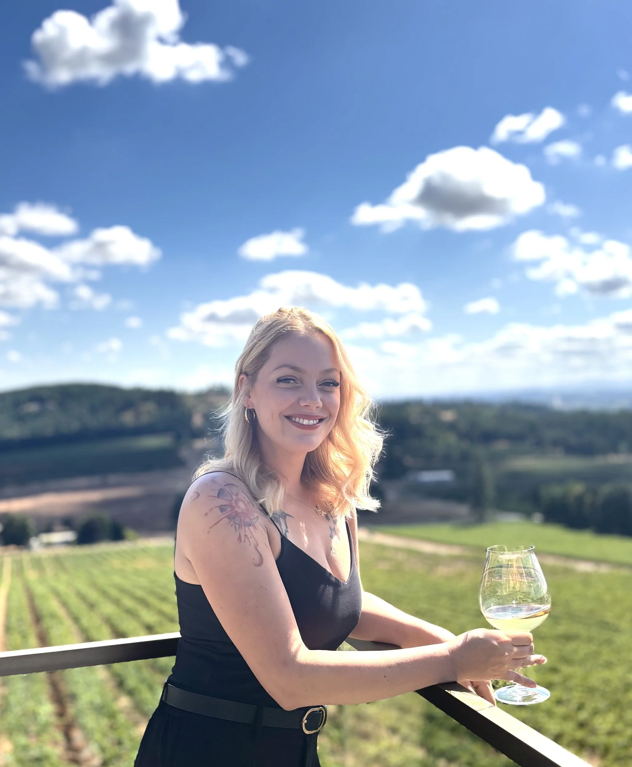 A woman with blonde hair and tattoos on her arm, wearing a black dress, is holding a wine glass filled with white wine while smiling outdoors on a balcony with a scenic view of fields and trees under a partly cloudy sky.