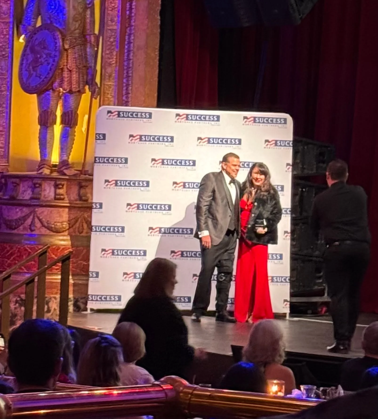 A man and woman posing together on stage at a success event, with a backdrop displaying multiple 'SUCCESS' logos. They are smiling, with the man in a suit and the woman in a red dress. Audience members are visible in the foreground, and a decorative 