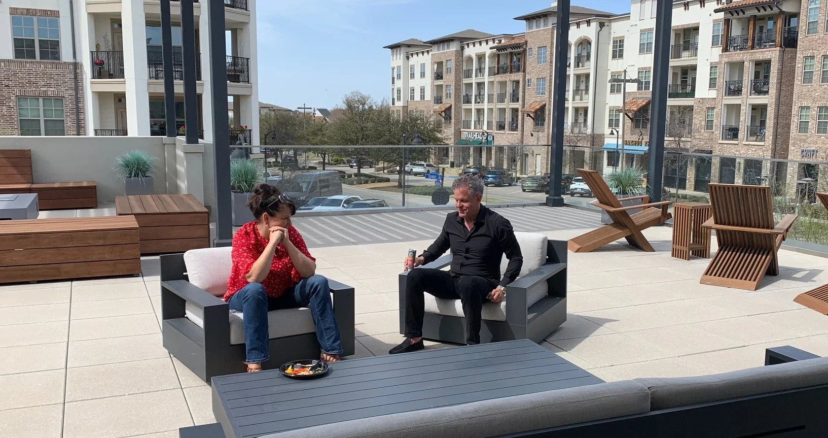 Two people sitting on outdoor couches on a patio, engaged in conversation. One is a woman with short dark hair, wearing a red polka dot blouse and jeans. The other is a man with gray hair, wearing a black shirt and black pants, holding a soda can. There are wooden chairs and plants in the background, with a glass railing and apartment buildings visible beyond.