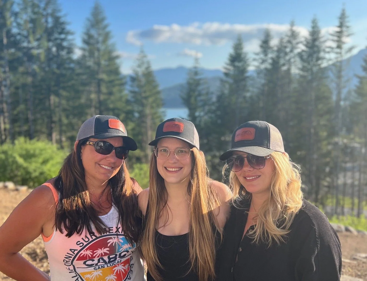 Three women wearing black and gray caps with red patches, sunglasses, and casual clothing, smiling outdoors with a forested mountain landscape in the background.