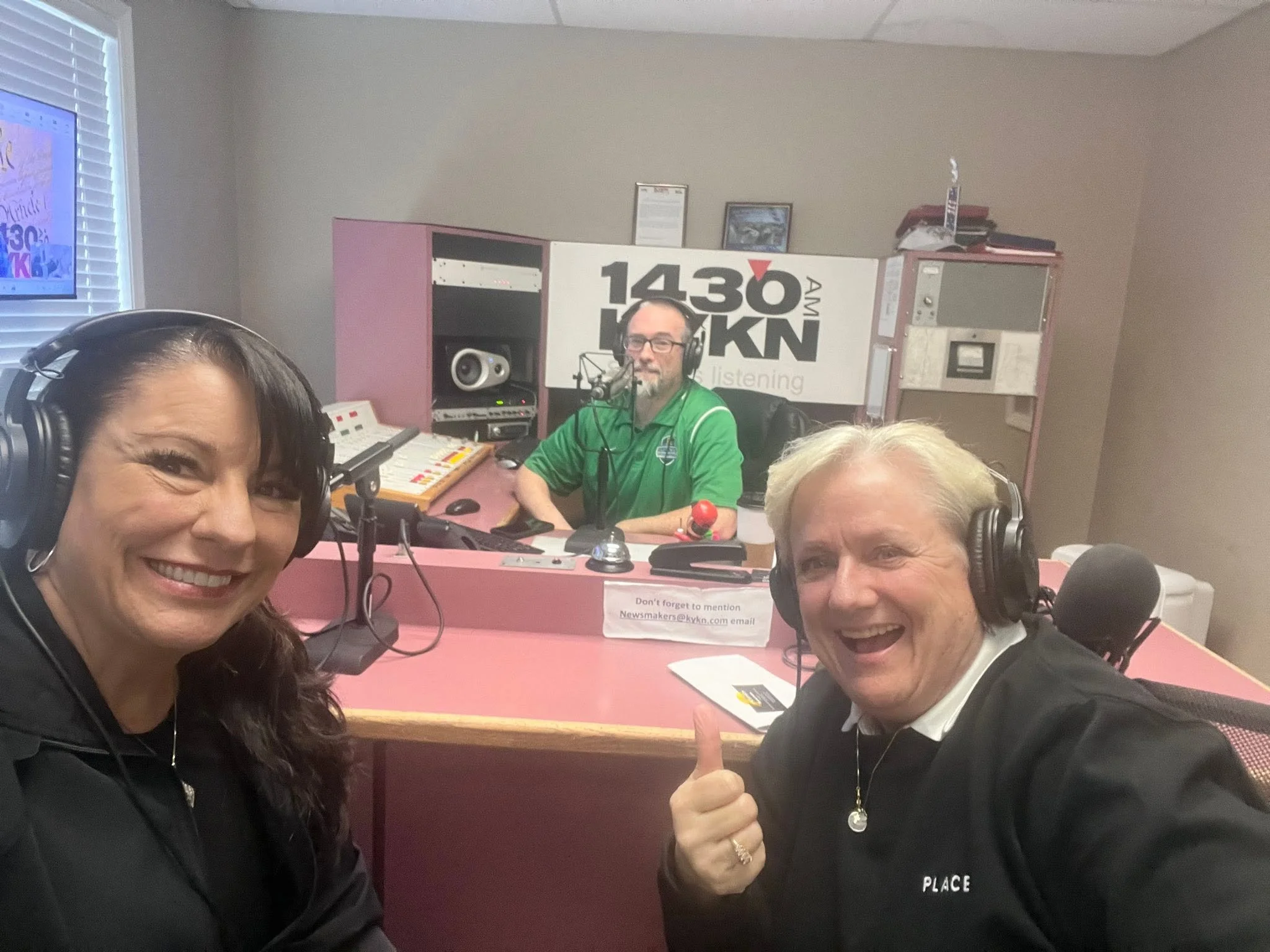 Four people in a radio studio, two women and two men, all wearing headphones and smiling. One woman is in the foreground, and the man is giving a thumbs-up. Behind them are radio equipment, a large sign for 1430 AM and 102.1 FM KKN, and a window with