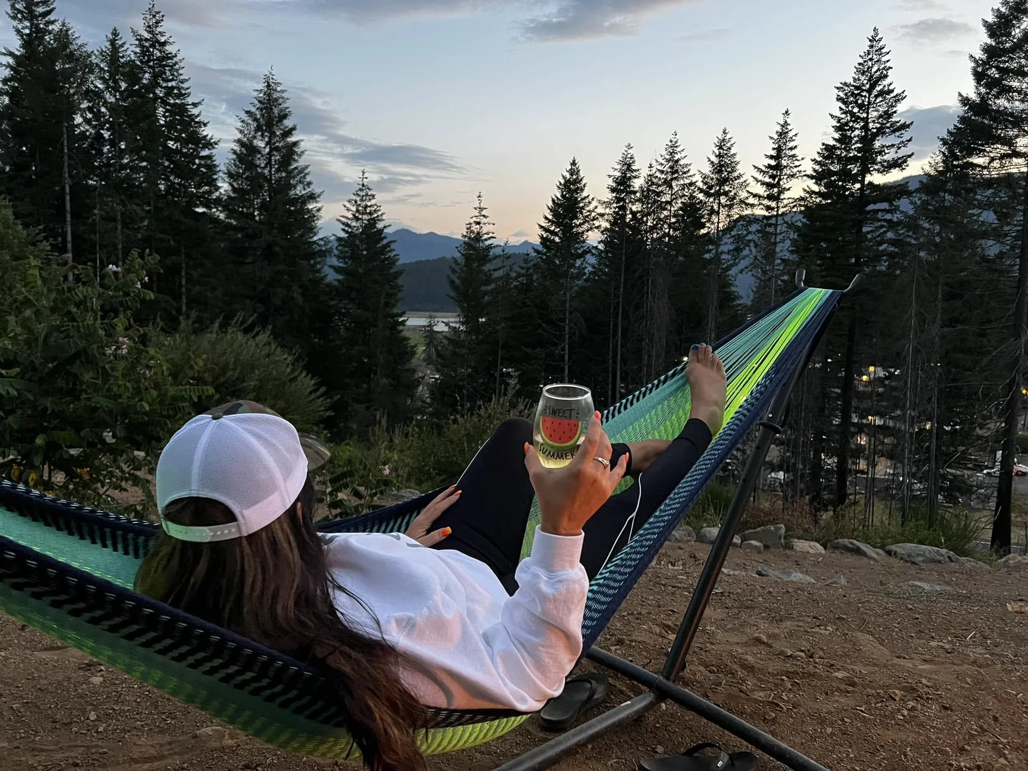 Person relaxing in a hammock at sunset, holding a glass of watermelon-themed drink, with a mountain landscape in the background and trees surrounding the area.