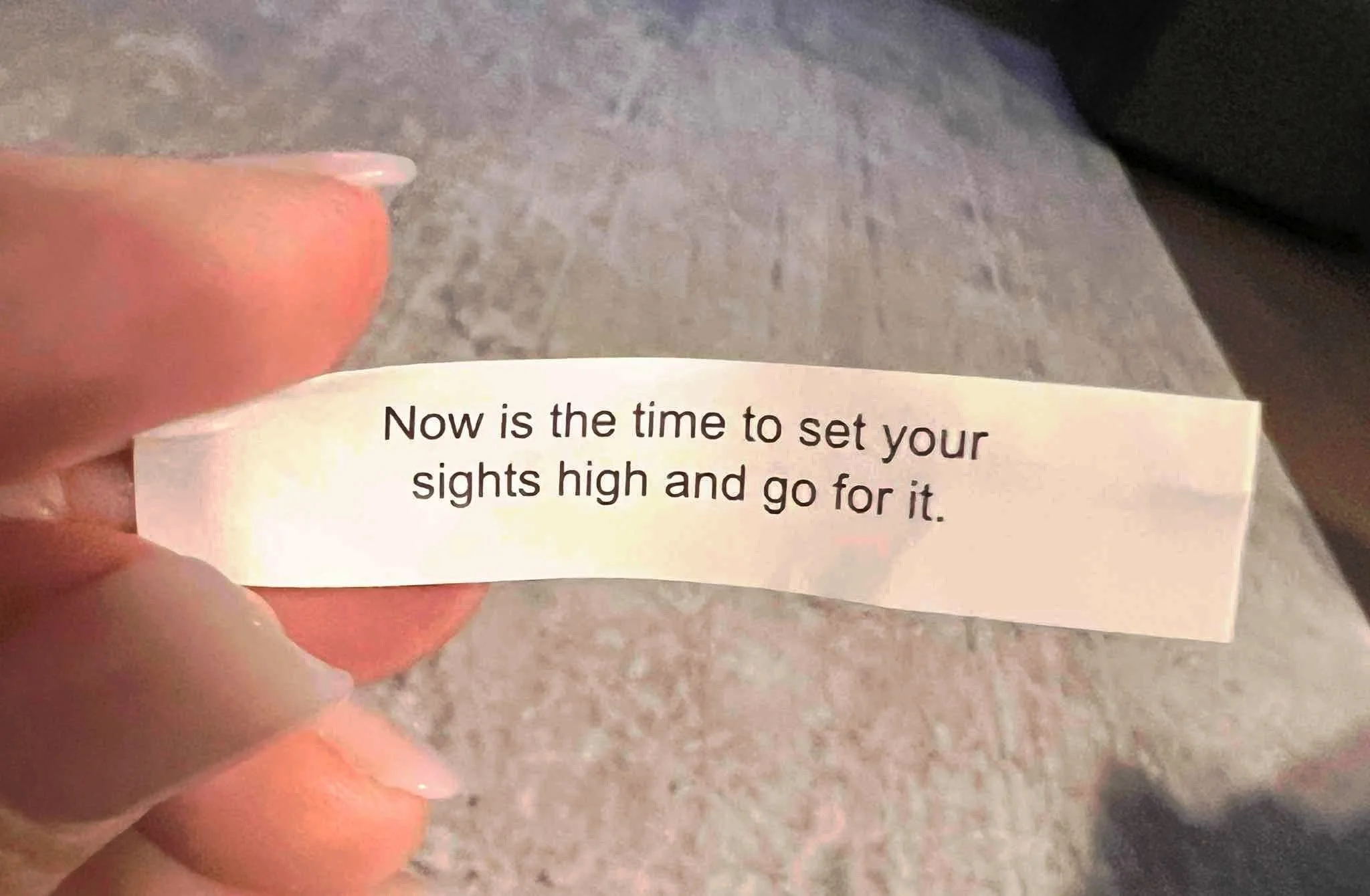 Close-up of a person's hand holding a piece of paper with the text: 'Now is the time to set your sights high and go for it.'