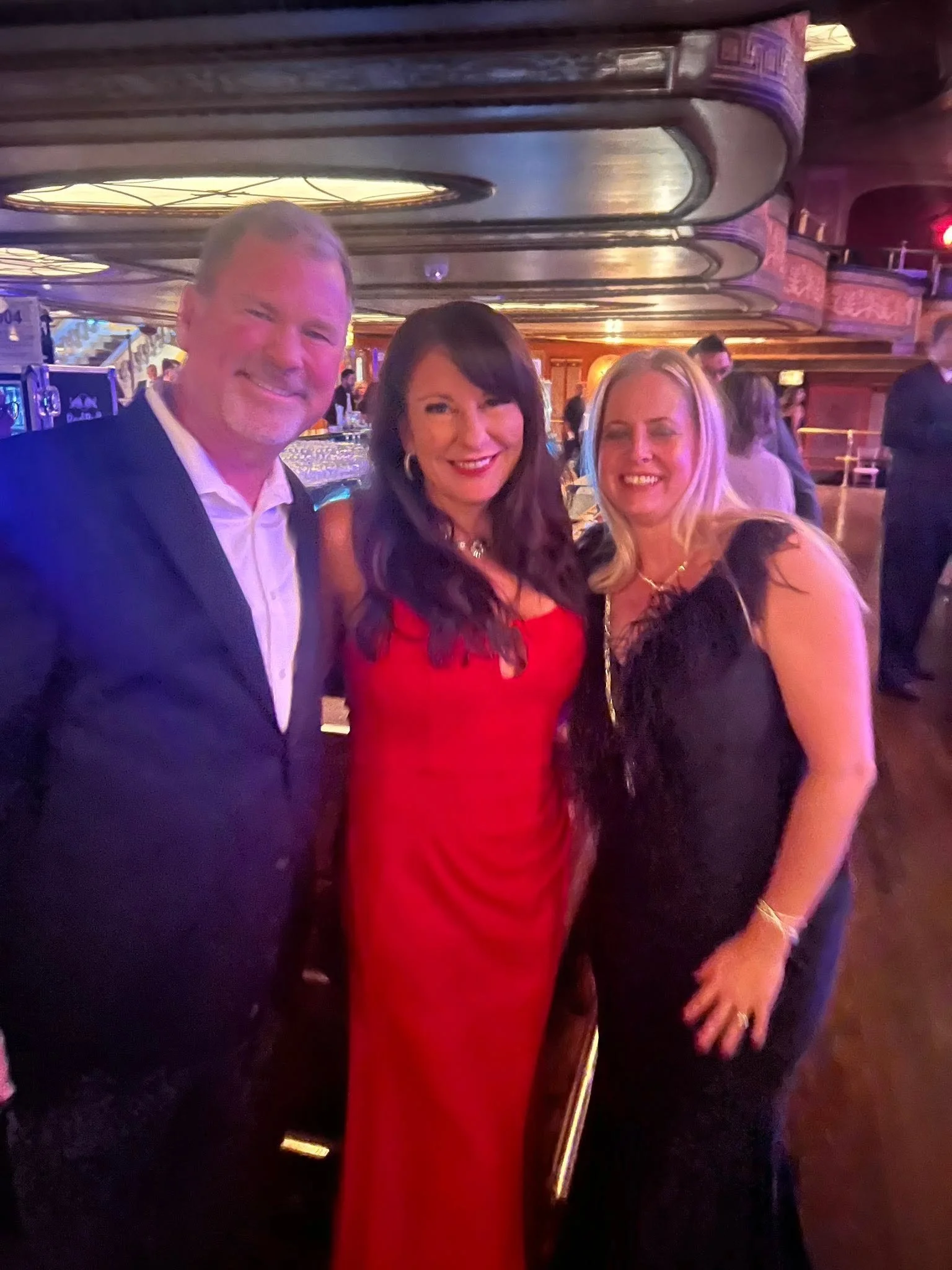 Three people dressed formally, two women and a man, smiling and posing for a photo in a dimly lit, elegant venue with a shiny ceiling and other people in the background.