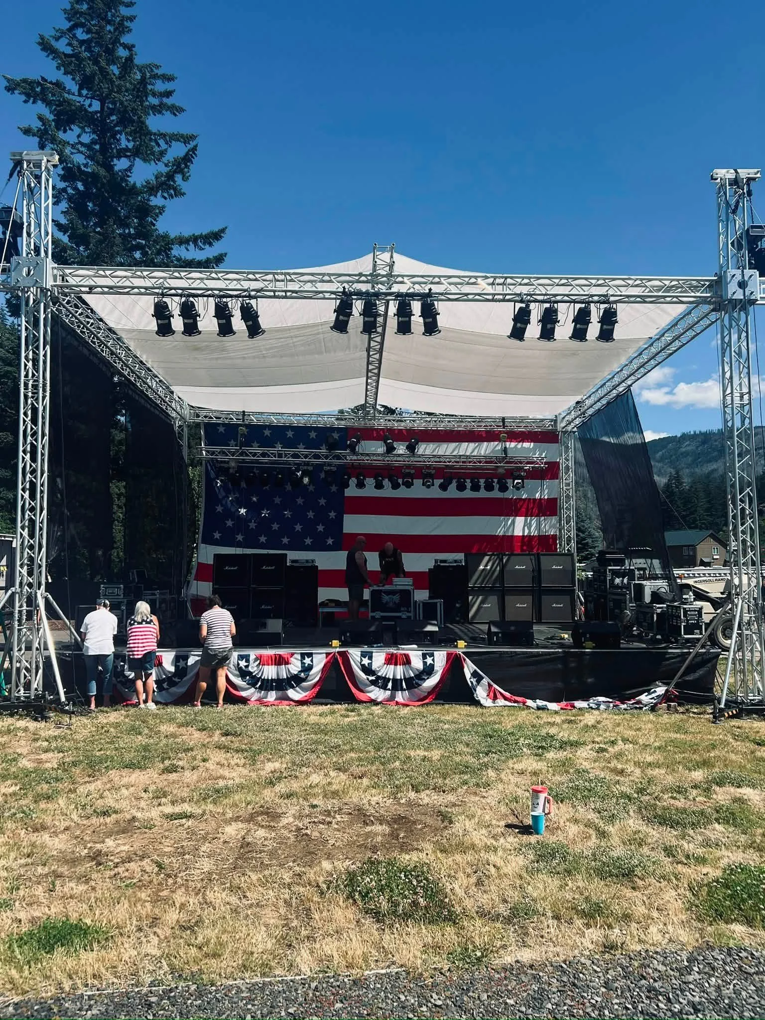 An outdoor stage decorated with an American flag theme, set up in a grassy area with a mountain and trees in the background, and some people preparing for an event.