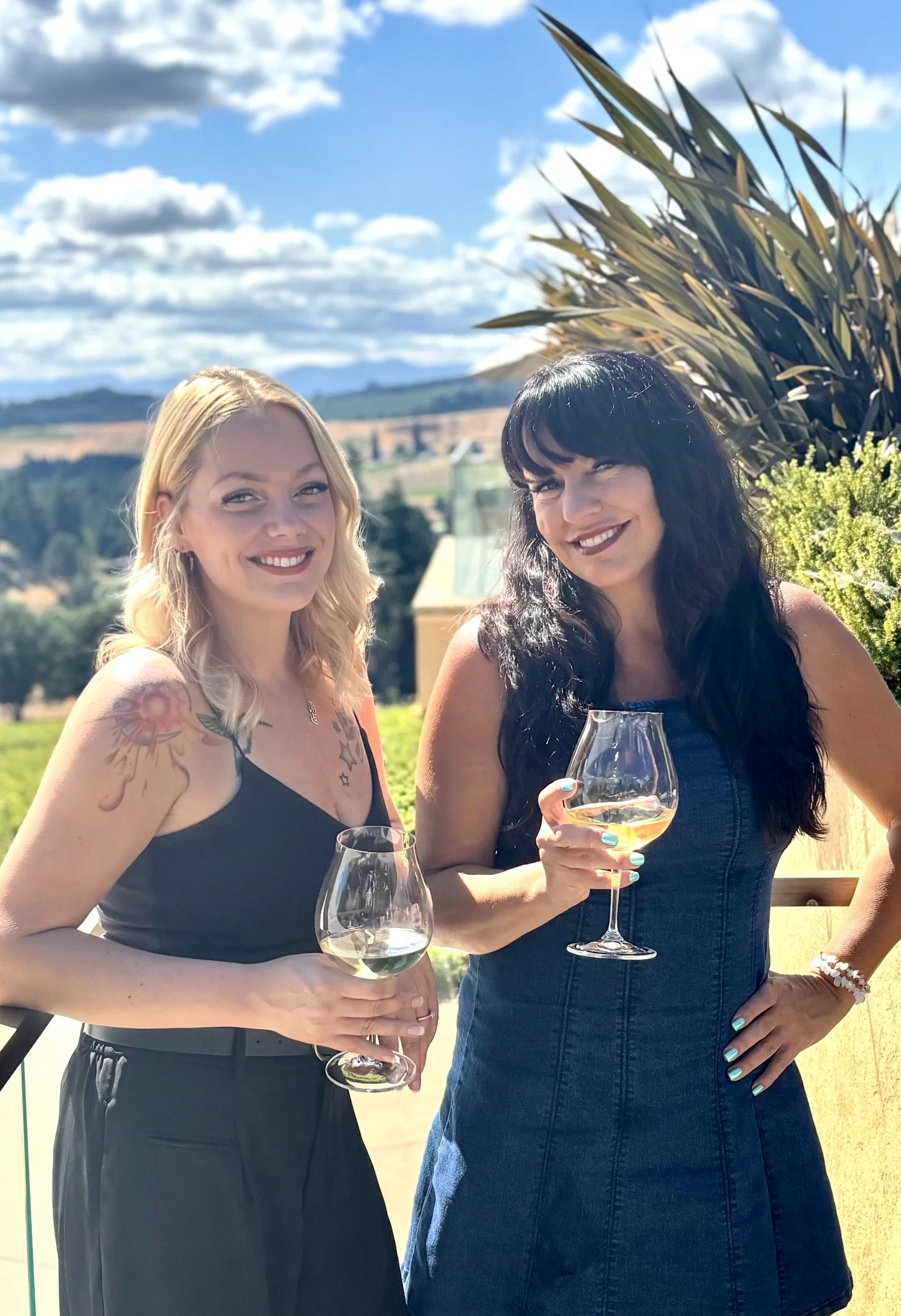 Two women smiling and holding glasses of white wine outdoors on a sunny day, with a blue sky and puffy clouds in the background.