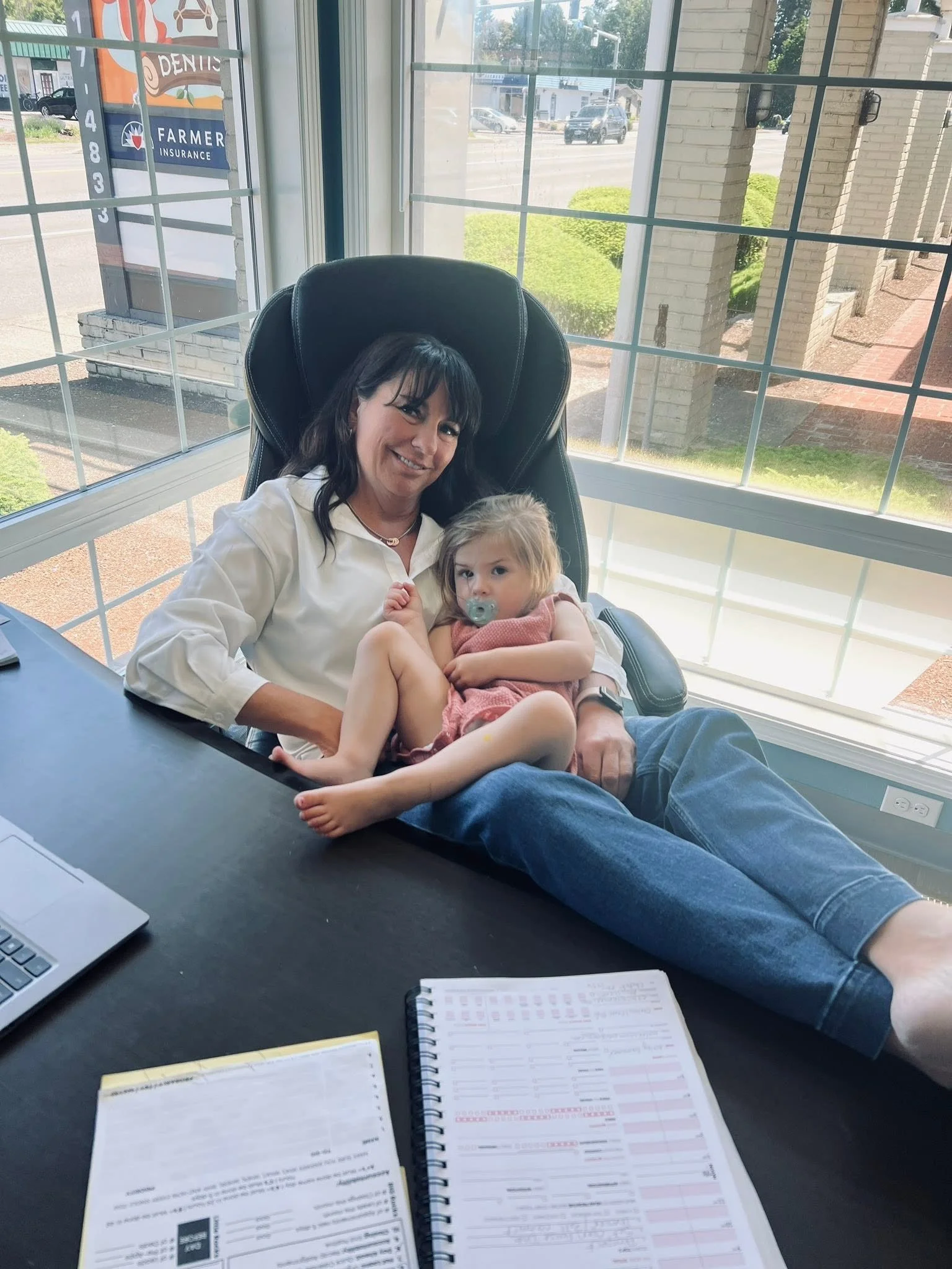 A woman and a young girl sitting in an office with large windows, the woman smiling and the girl holding a pacifier, with office papers and notebook on the desk.