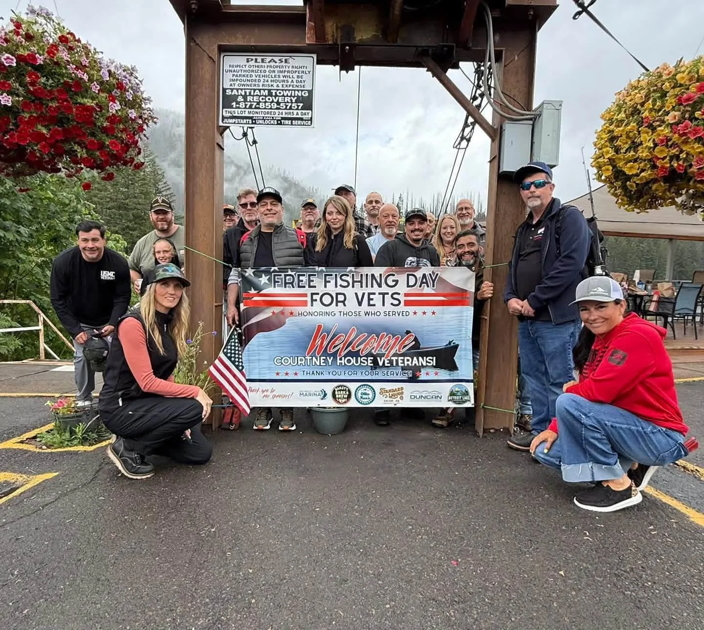 Group of people holding a banner that reads 'Free Fishing Day for Vets' in front of a wooden structure. Some individuals are kneeling or crouching in front, others are standing behind the banner. The setting appears to be outdoors near a marina or fi