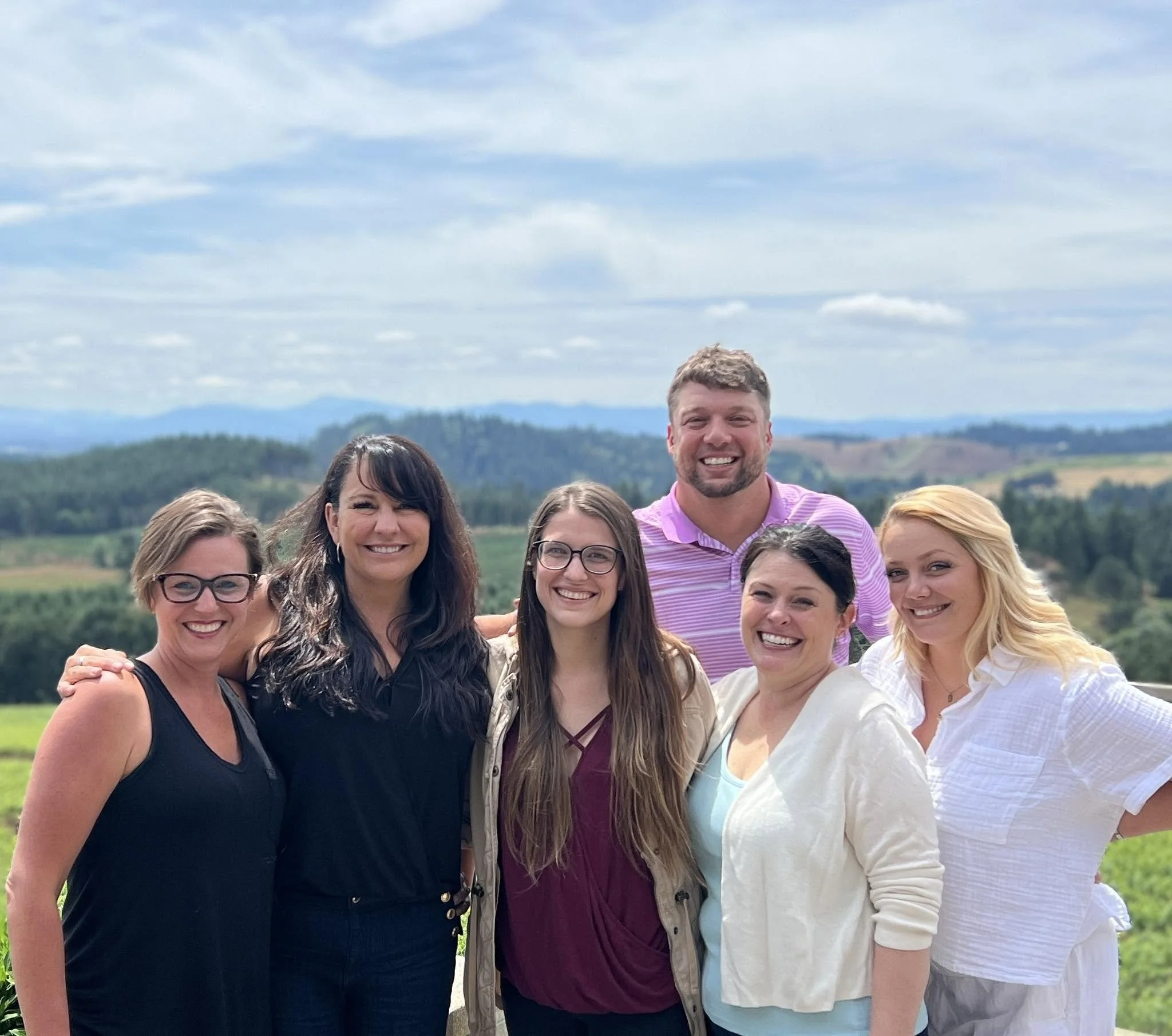 A group of smiling women and one man standing outdoors with a scenic landscape of rolling hills and a cloudy sky in the background.