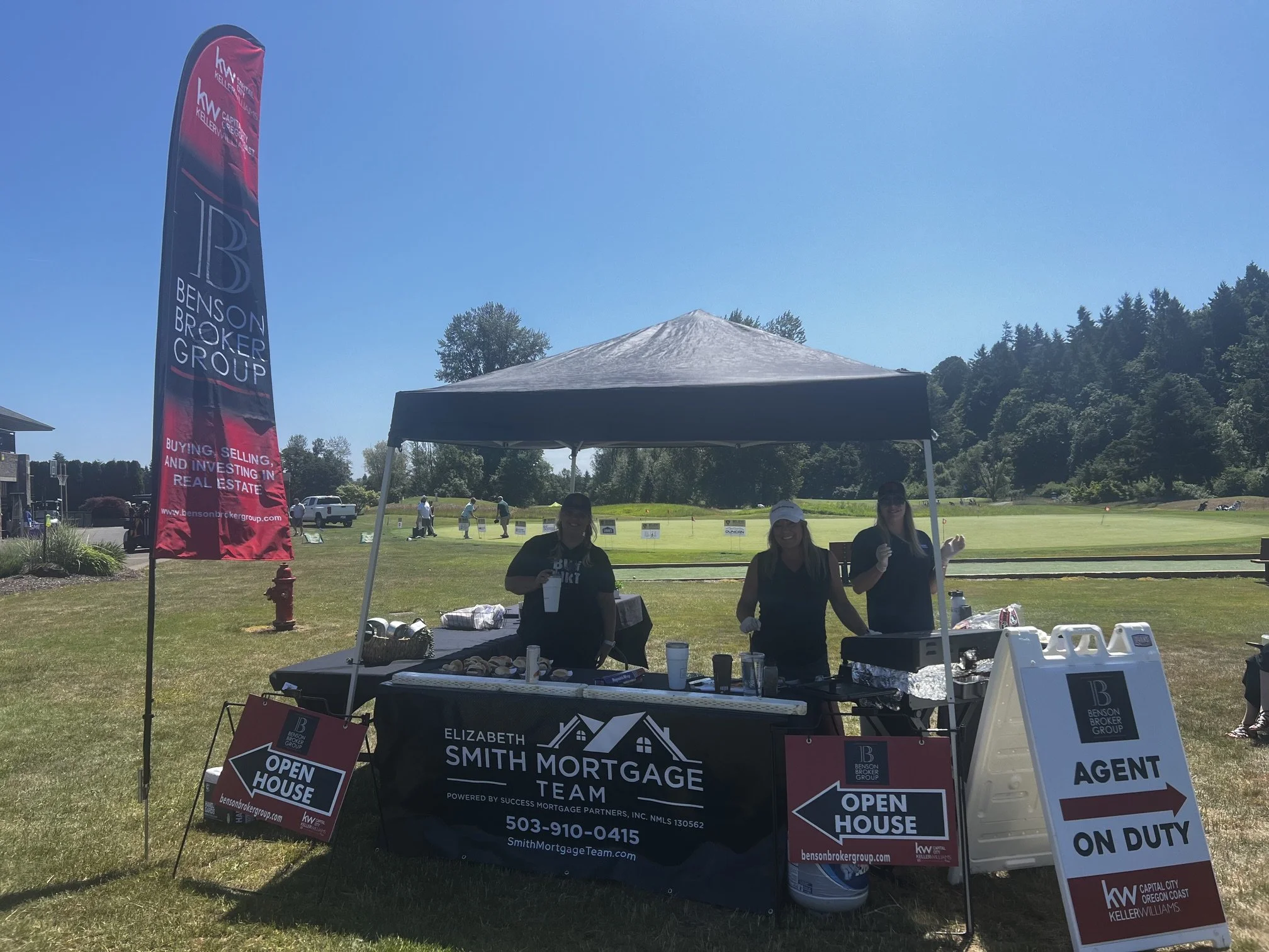 Outdoor real estate event with banners and signs for Smith Mortgage Team, open house, and on-duty agent. Three women smiling near the tables under a canopy, with a golf course and trees in the background.
