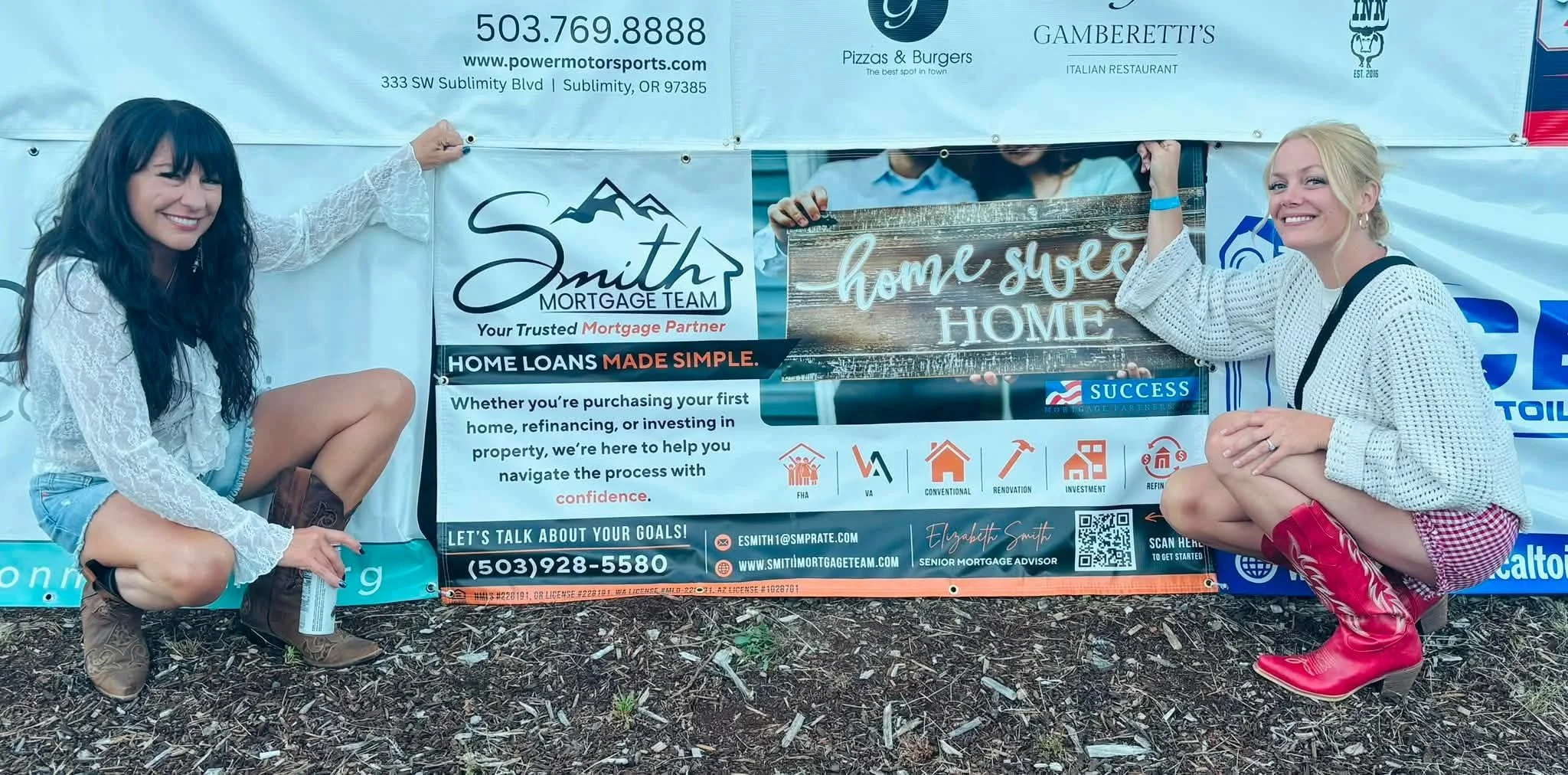 Two women crouch on either side of a large banner at an outdoor event, smiling at the camera. The banner displays advertisements for mortgage services, including contact information, logos, and slogans like 'home sweet home' and 'home loans made simp