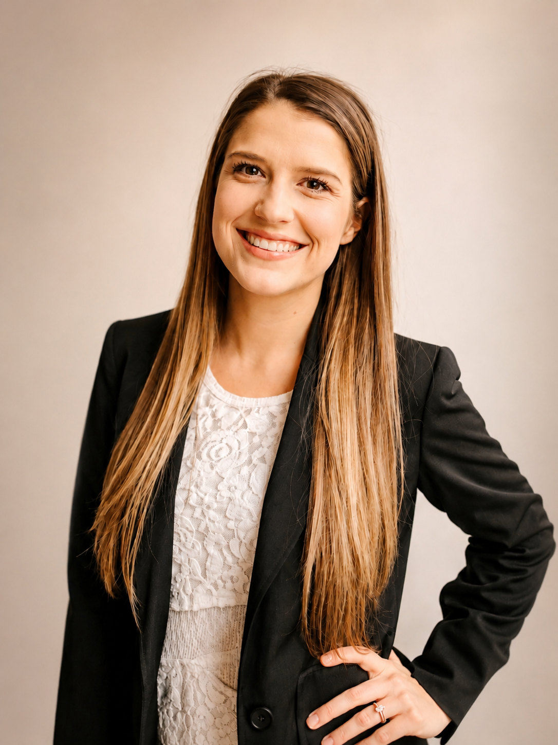 A woman with long brown hair smiling, standing with her hand on her hip against a neutral background, wearing a black blazer over a white lace top, and a wedding ring on her finger.