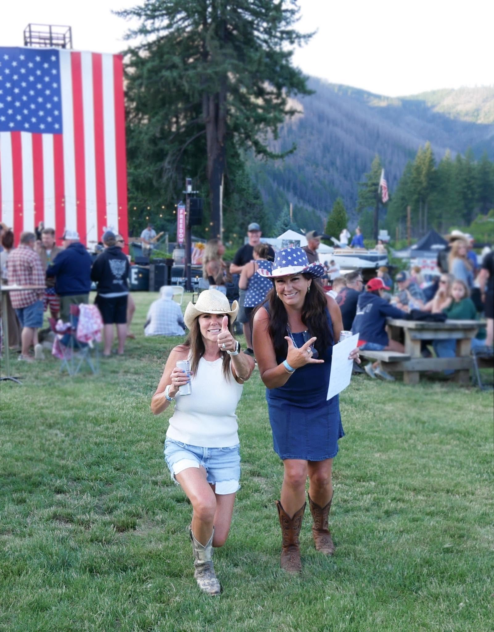 Two women smiling and walking on grass at an outdoor event; one wearing a cowboy hat, the other in a patriotic hat, with a crowd and an American flag backdrop in the background.