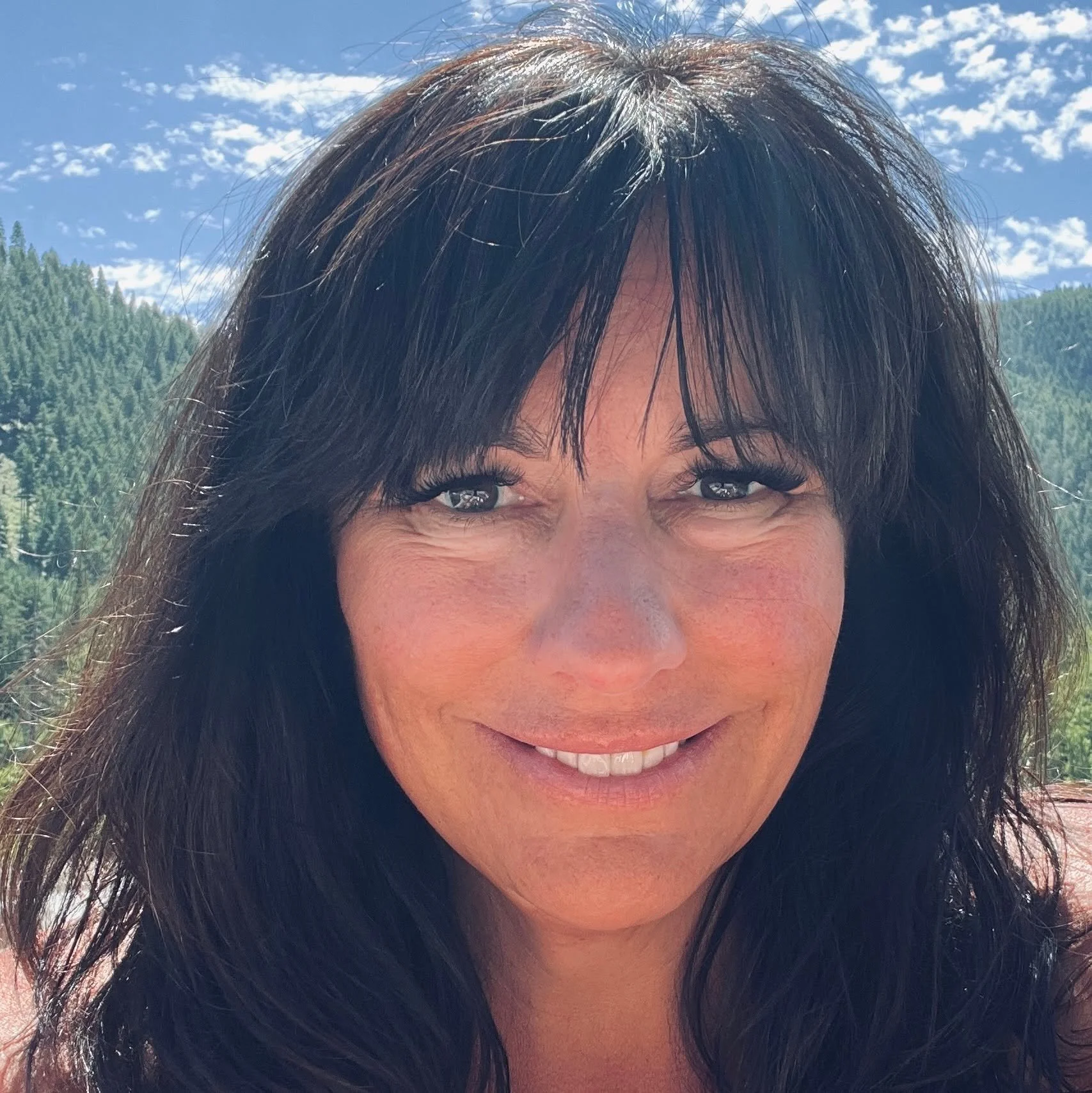 Close-up of a woman's face with dark hair, smiling outdoors under a blue sky with scattered clouds and a green forest background.