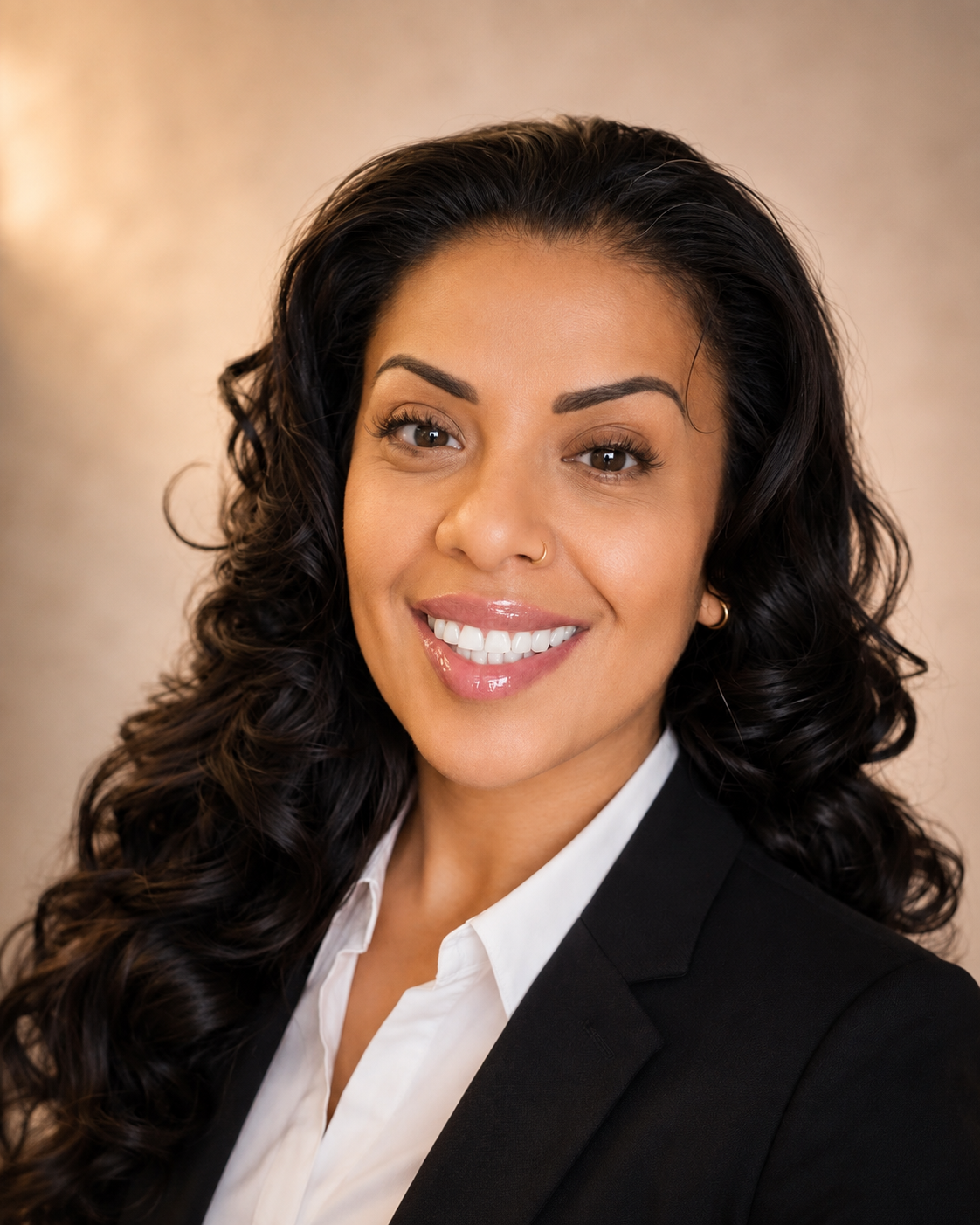 A professional woman with dark, curly hair smiling, wearing a black blazer and white shirt, with a beige background.