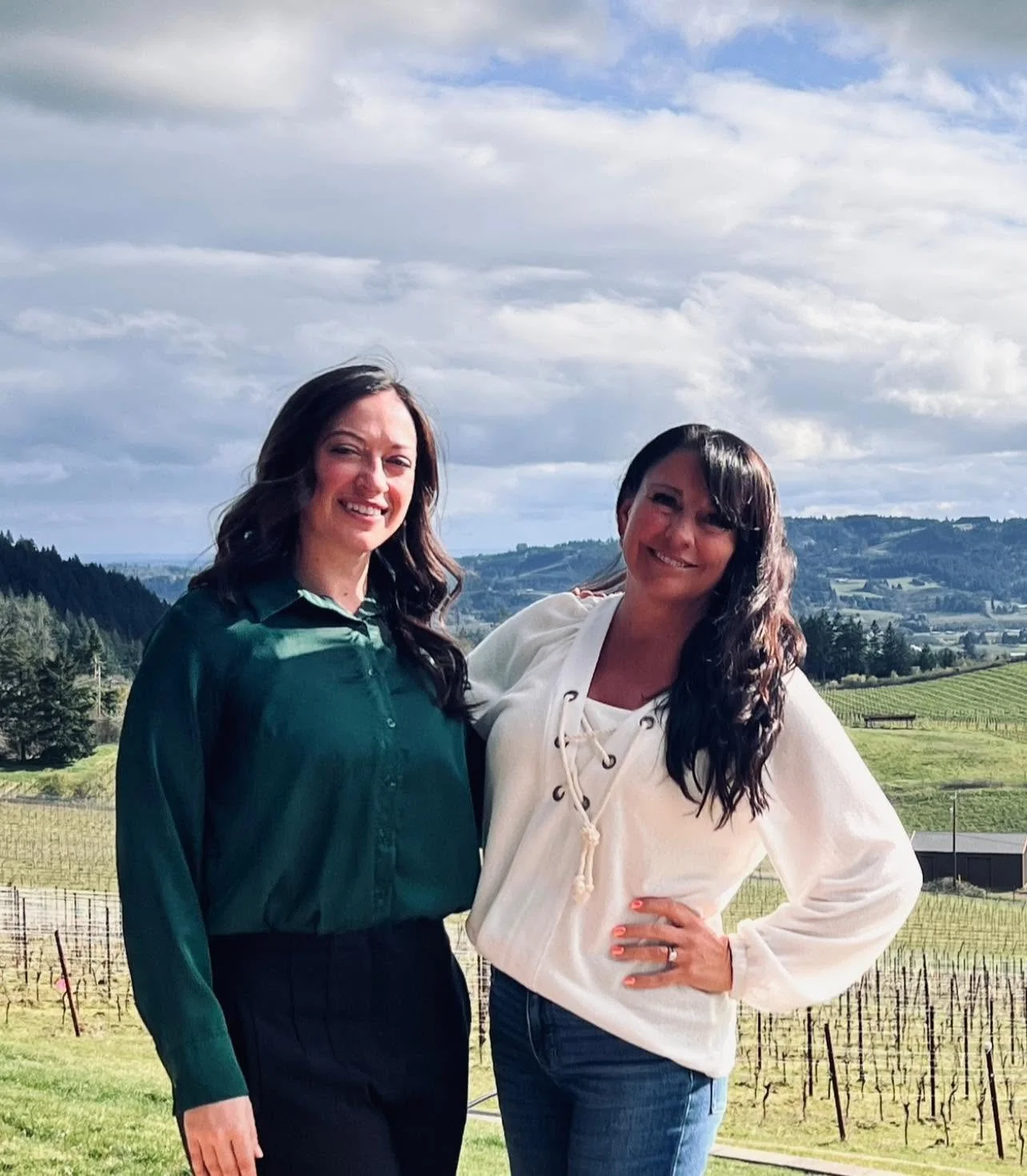 Two women standing outdoors in a vineyard with rolling hills and a cloudy sky in the background, smiling at the camera.