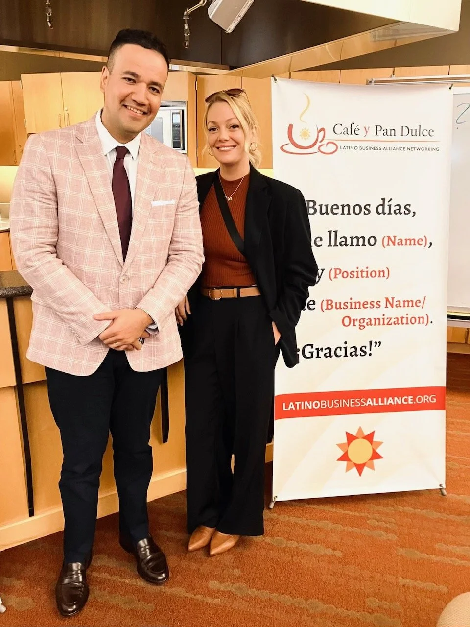 A man and woman standing together indoors, smiling at the camera, near a sign that reads 'Café y Pan Dulce' and promotes Latin American business networking.