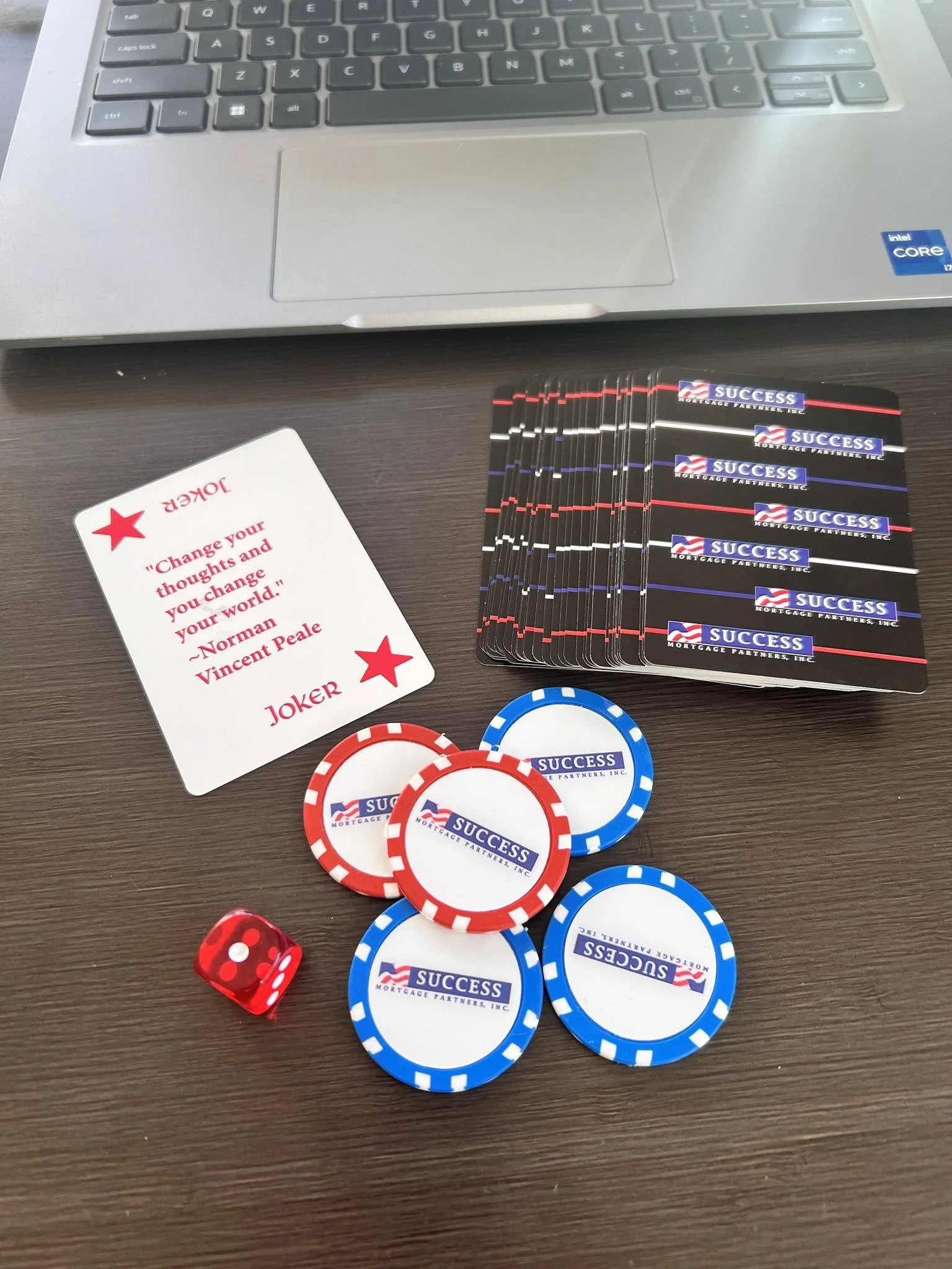 A laptop keyboard, a playing card with a quote, a red die, poker chips with 'SUCCESS' and 'MORTGAGE PARTNERS, INC.' printed on them, and some red and black gaming chips on a dark wooden surface.