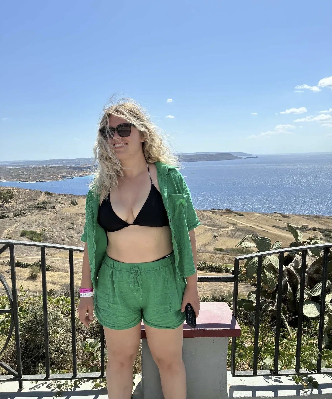 A woman with blonde curly hair wearing sunglasses, a black bikini top, a green shirt, and green shorts, standing outdoors on a sunny day near water and a hillside with sparse vegetation.