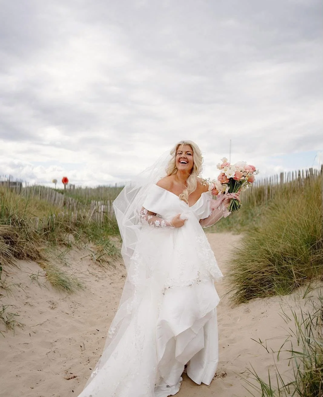 A bride in a white wedding dress holding a bouquet of pink and white flowers walking on a sandy beach path with grass and dunes, under a cloudy sky.