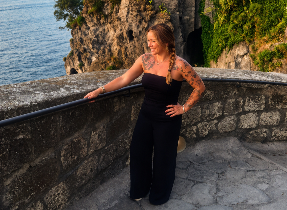 A woman with tattoos on her arms and shoulder, wearing a black strapless top and black pants, stands on a stone walkway by the water, smiling and looking to her right, with a rocky cliff and green foliage in the background.