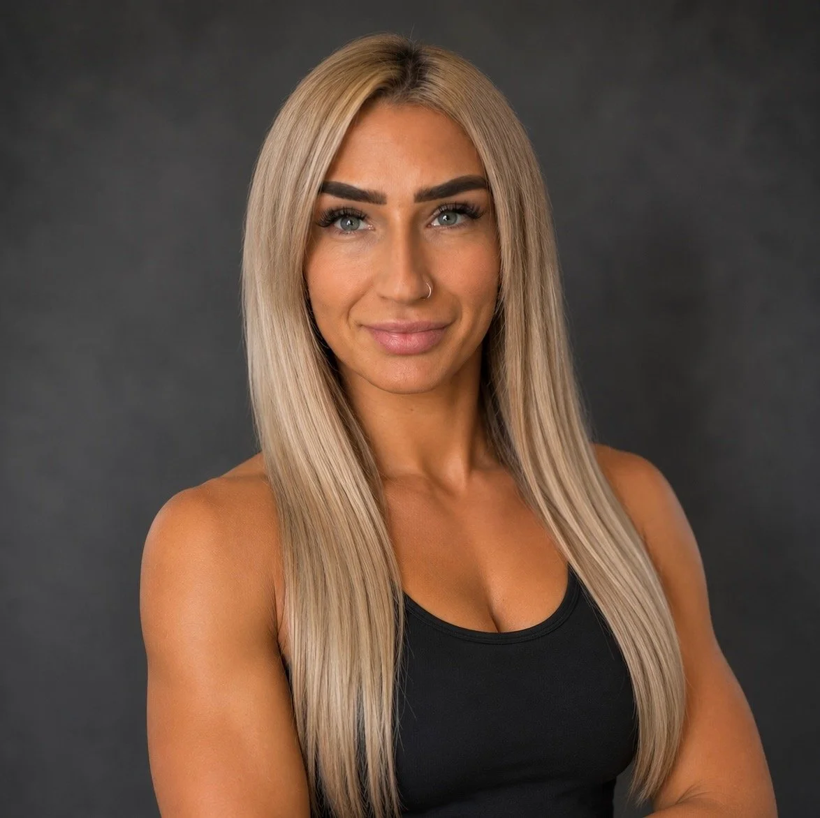 A woman with long blonde hair, blue eyes, wearing a black sleeveless top, smiling with a confident expression, against a dark gray background.