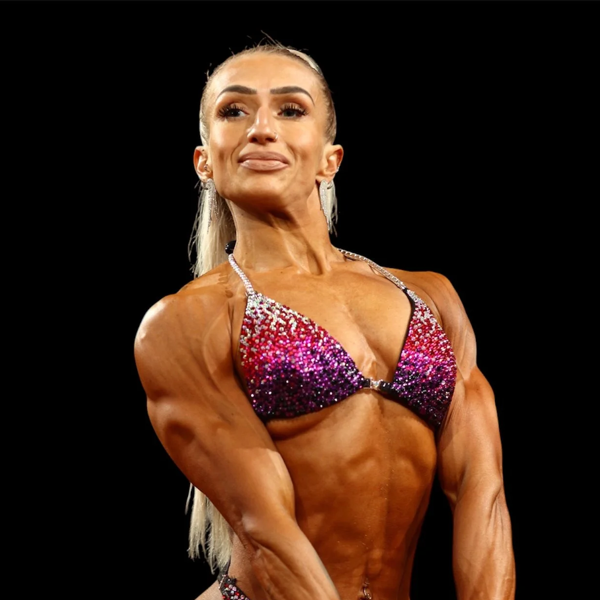 A female bodybuilder in a glittery bikini posing on stage with a black background.