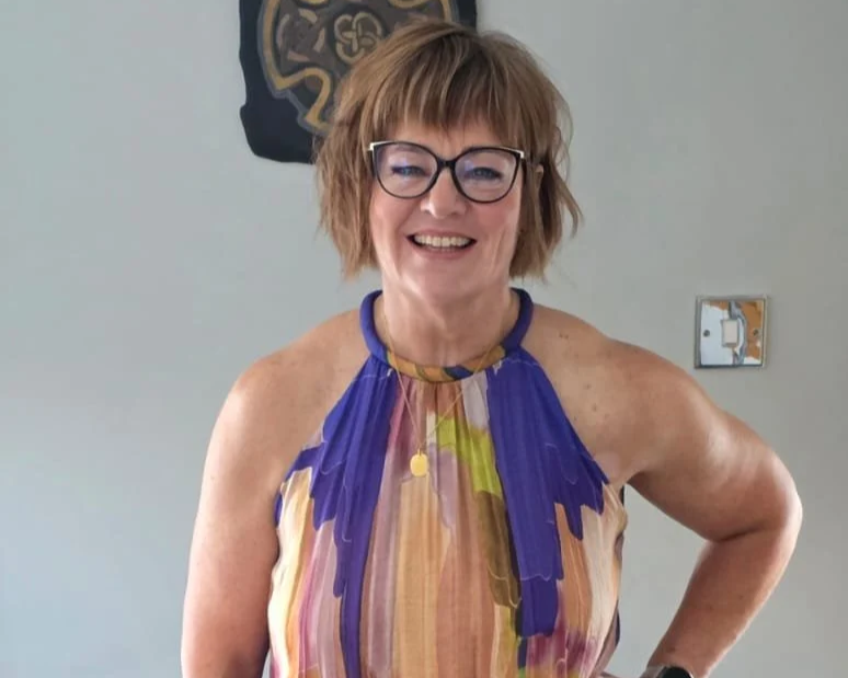 A woman with short, messy brown hair and glasses, smiling, wearing a colorful striped halter top, standing indoors against a plain wall.