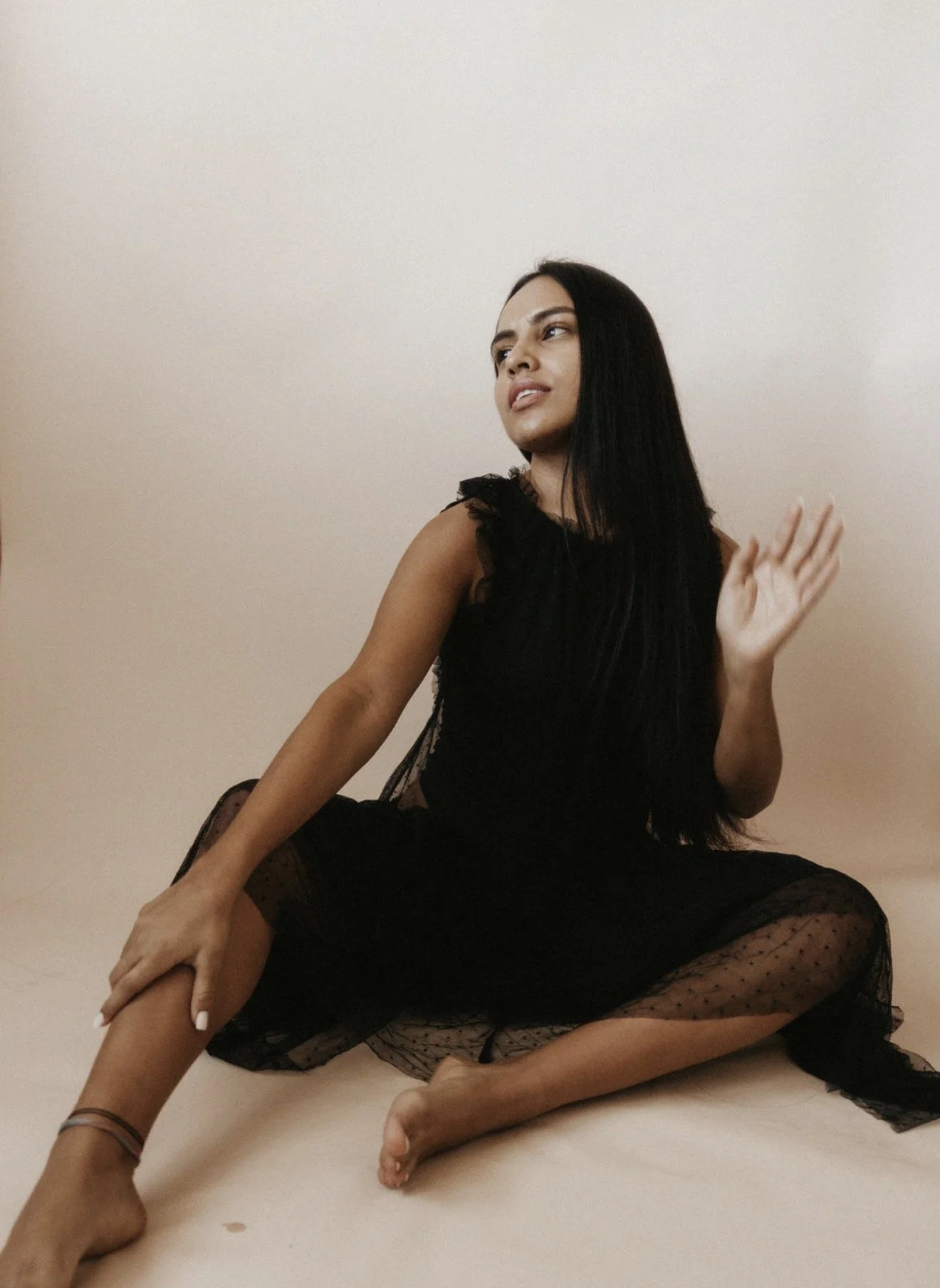 A woman with long black hair sitting on the floor against a beige wall, wearing a black dress with lace details, and posing with her head slightly tilted and a hand raised.