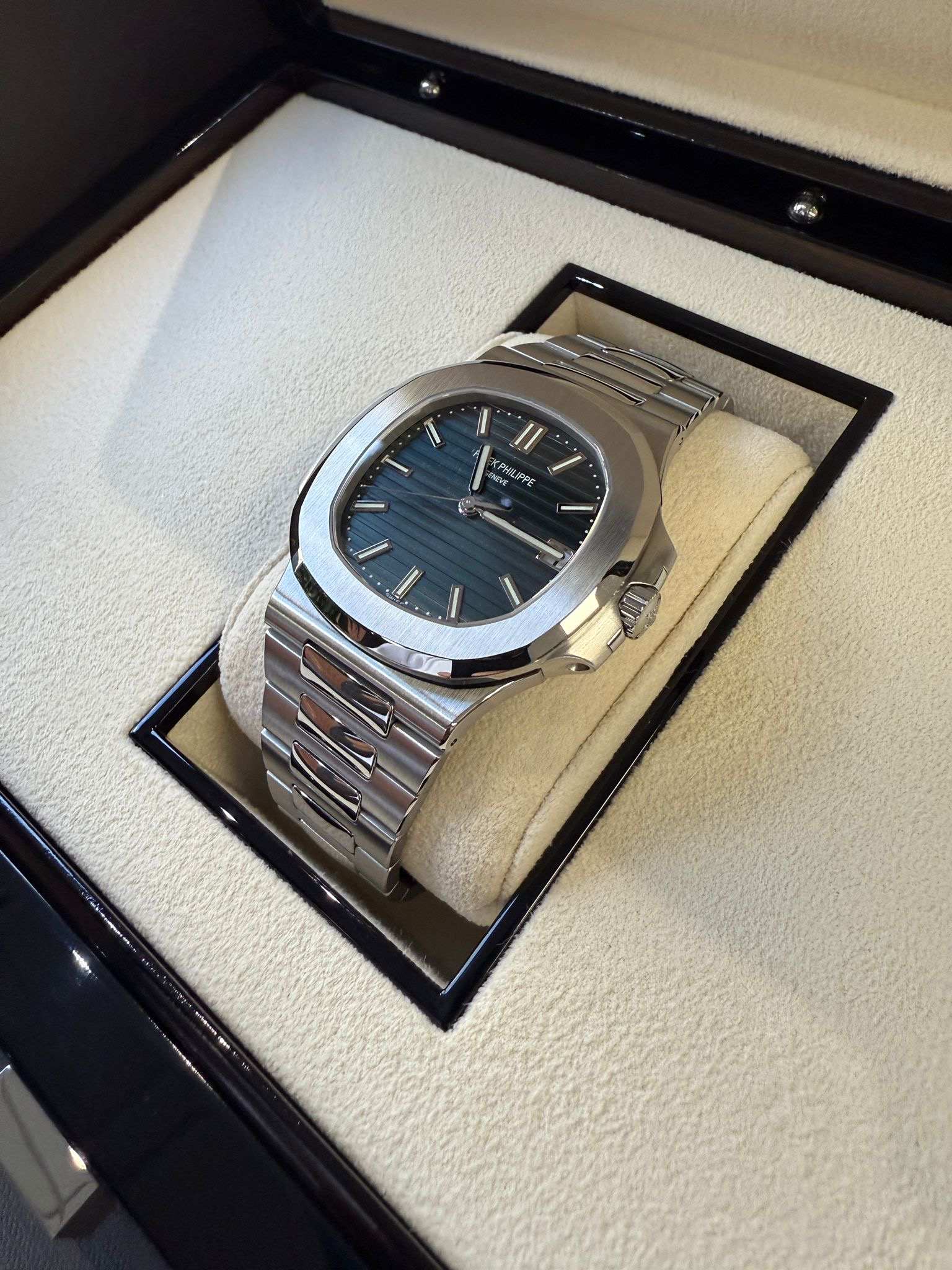 A luxury watch inside a black box with a beige interior, featuring a dark blue dial, silver markers, and a silver bracelet.