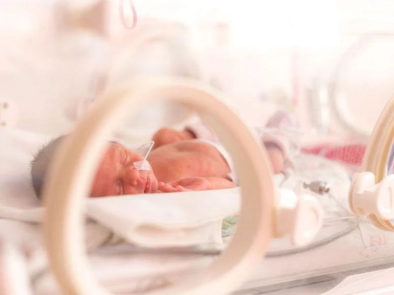 Newborn baby in a neonatal intensive care unit lying in a clear incubator, with a nasal feeding tube attached.