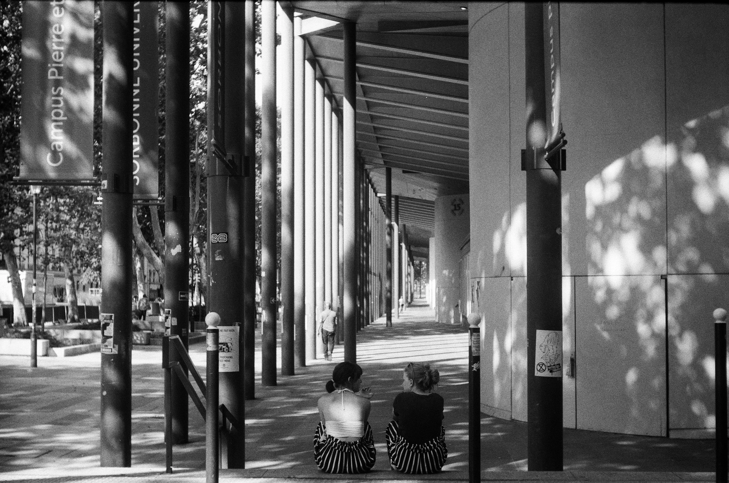 Two women sitting on the sidewalk in front of a modern building with vertical poles and trees in the background. They are facing away from the camera, engaged with their phones.