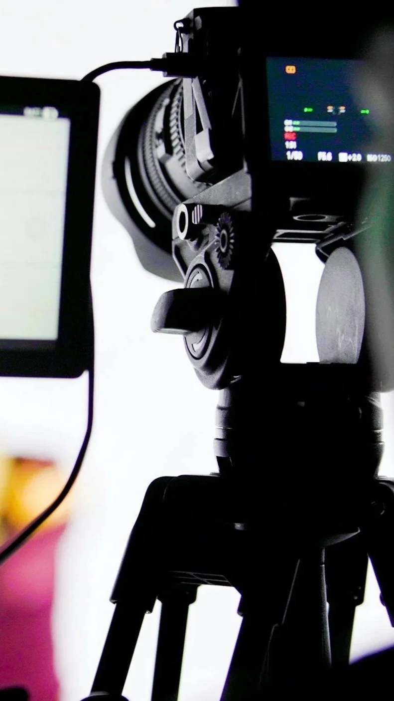 Close-up of a camera mounted on a tripod with a monitor screen showing camera settings, against a plain background. Self Tapes for Actors, acting coaching and tips.