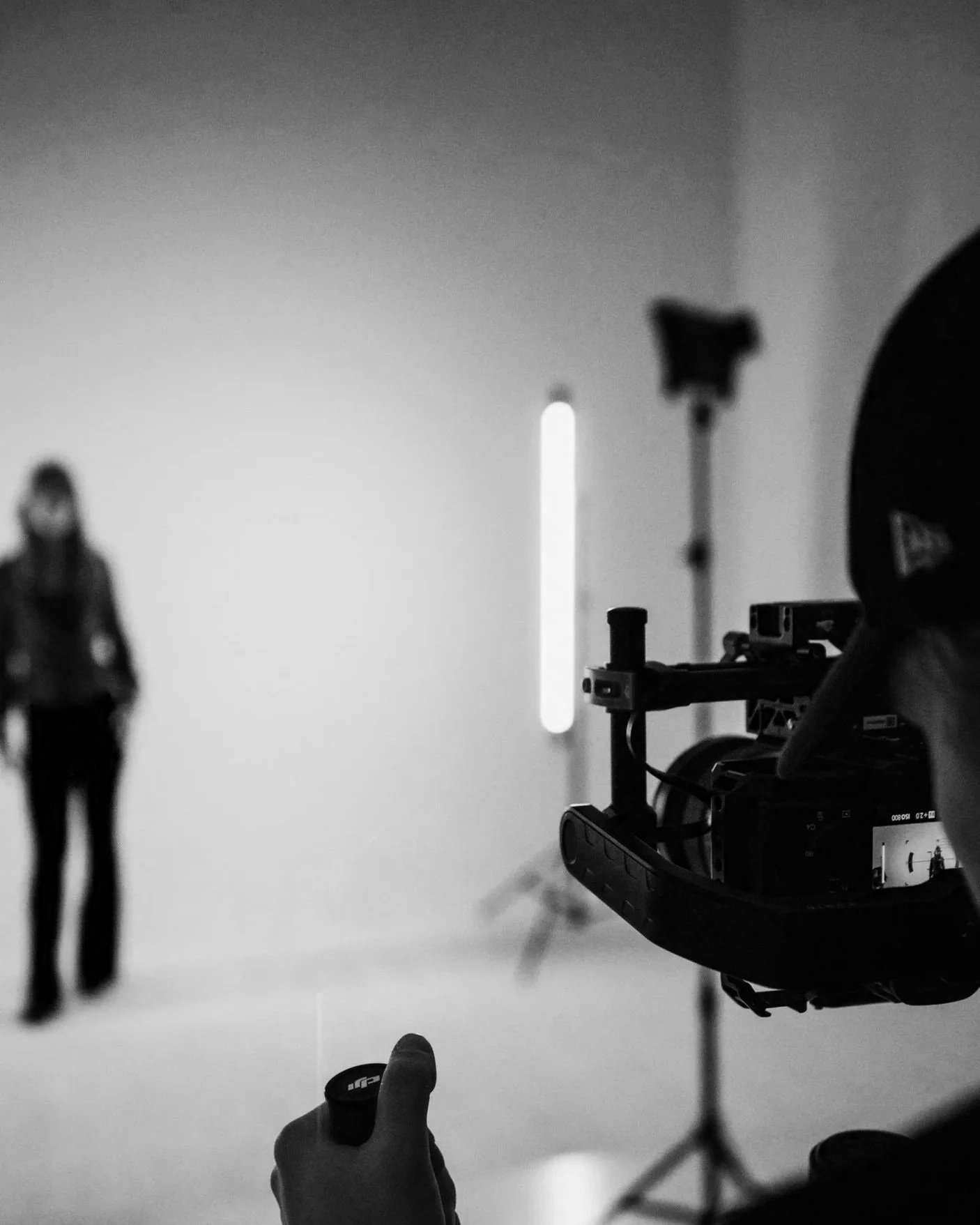 Black and white photo of a film or photography set with a camera on a tripod, a light, and a woman standing in the background, out of focus.