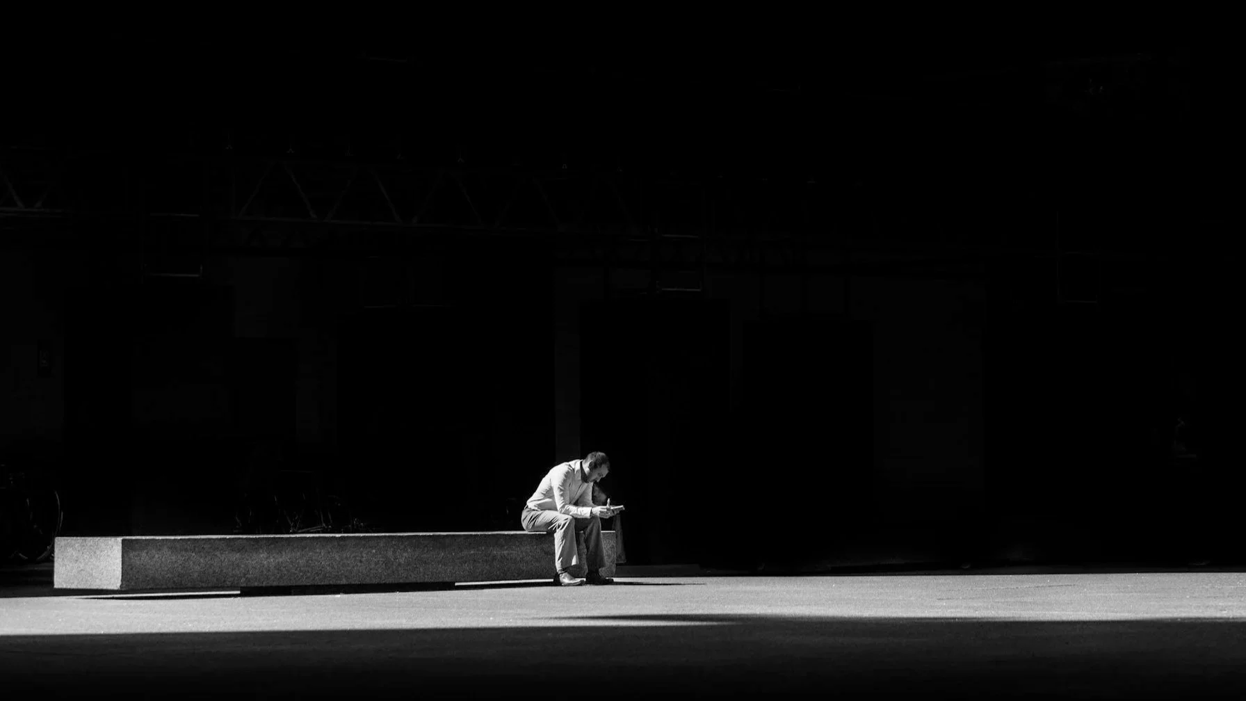 A man sitting on a bench, reading or looking at his phone, in a dark, urban setting with high contrast lighting.