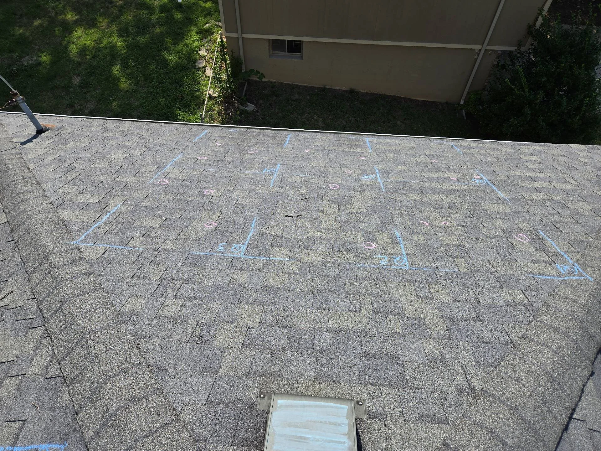 Roof with chalk markings for shingle repairs or replacements, in a residential area with a neighboring house and some greenery in the background.