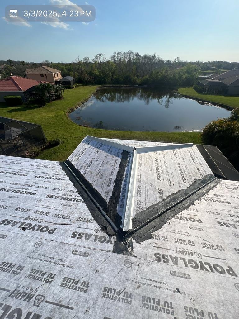 A rooftop with a skylight under construction, overlooking a backyard pond surrounded by green lawns and residential houses on a clear day.