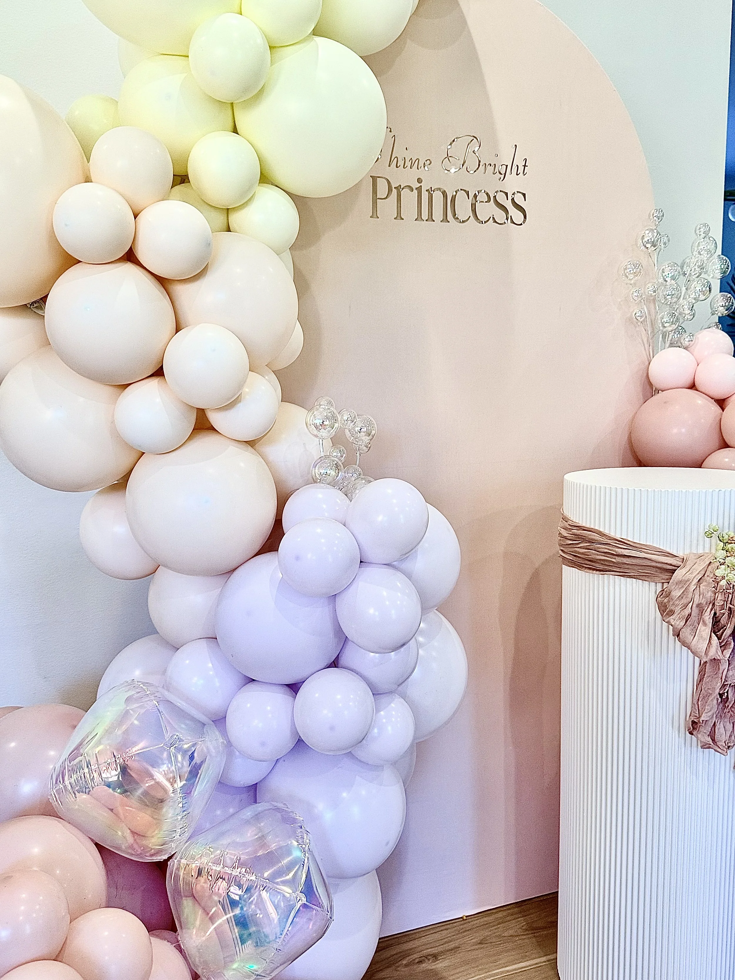 A decorative balloon arrangement with pastel pink, purple, white, and cream-colored balloons, along with transparent iridescent balloons, set against a pink wall. There is a sign that says "Shine Bright Princess" on the wall, and a white, vertically 