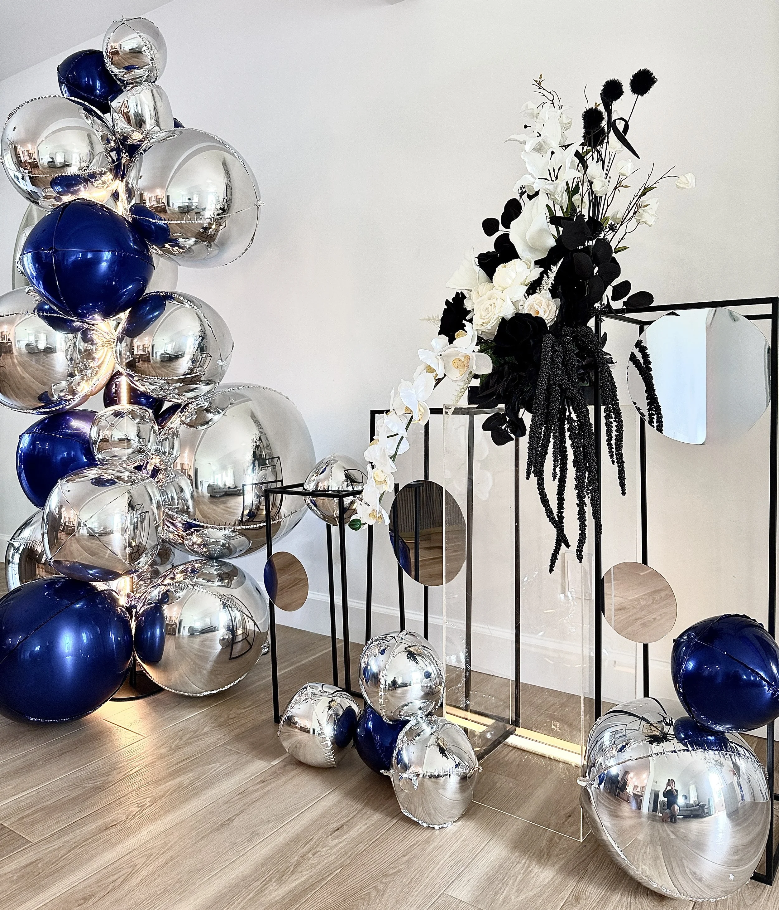 A modern celebration setup featuring reflective silver and deep navy balloons, layered acrylic and metal frames, mirrored accents, and a sculptural black-and-white floral arrangement. The asymmetrical layout creates a clean, contemporary focal point 