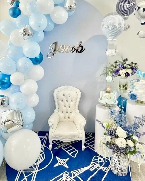 Decorative birthday display with blue and white balloons, a white tufted chair, and a cake table with purple flowers, a birthday cake, and balloons, with a sign reading 'Jacob' on the wall.