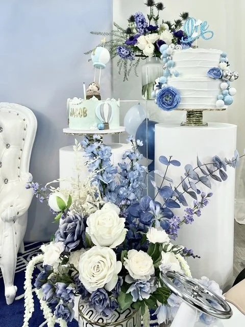 Elegant wedding or celebration cake display with white and blue flowers, including roses and hydrangeas, arranged on white pedestals and a table, featuring a multi-tiered cake decorated with blue and white accents and a floral arrangement in the back