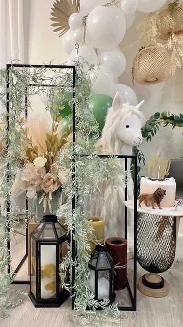 Decorative display with white balloons, gold woven wall art, a white plush unicorn, a small animal sculpture, floral arrangements, and candles in black lanterns.