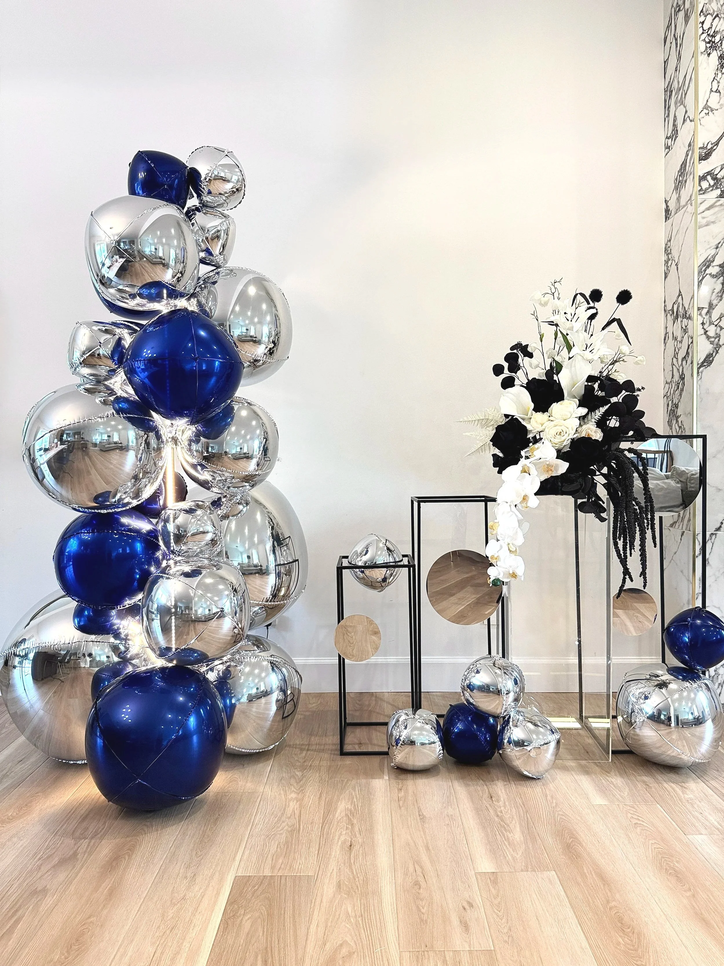 A modern celebration setup featuring reflective silver and deep navy balloons, layered acrylic and metal frames, mirrored accents, and a sculptural black-and-white floral arrangement. The asymmetrical layout creates a clean, contemporary focal point 