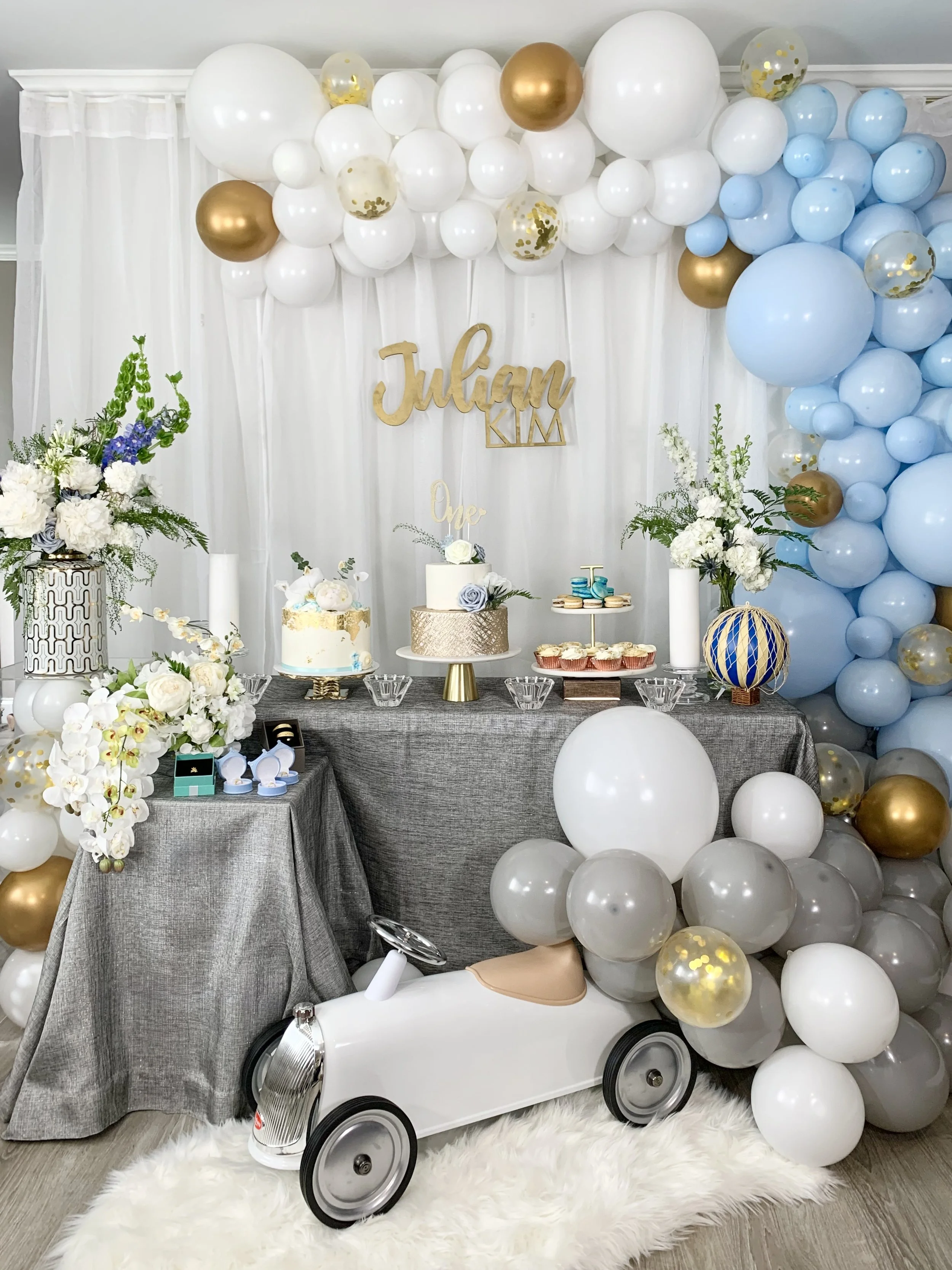 A decorated celebration area with white, gold, and light blue balloons, floral arrangements, and a cake table. There is a white balloon arch and a white toy car ride-on toy in the foreground with a fluffy white rug underneath.