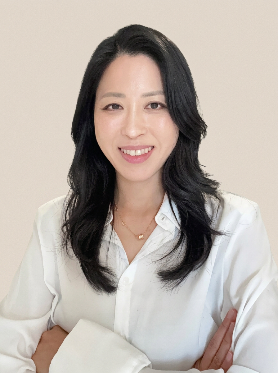A woman with long black hair, wearing a white blouse and a gold necklace, smiling and crossing her arms in front of a plain light-colored wall.