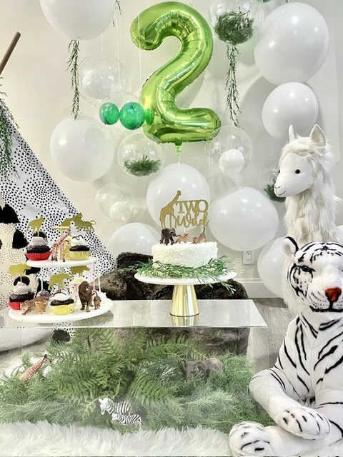 Decorated birthday celebration with white balloons, a green number 2 balloon, plush tiger and unicorn toys, and cupcakes with animal toppers, in a room with greenery and a stuffed zebra.