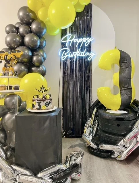 Decorations for a birthday party including black, yellow, and silver balloons, a cake with black, yellow, and white decorations, a neon sign that says 'Happy Birthday', and a large yellow and black number '3' balloon.